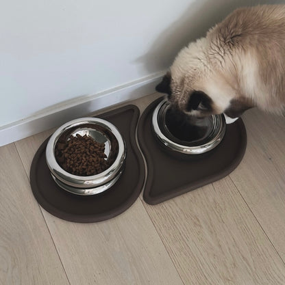 Maskad katt som äter från en matplats med två mörkbruna espressorunfärgade silikonunderlägg på ett ljust trägolv
