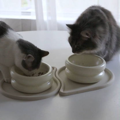 En gråvit katt och en långhårig grå katt som äter tillsammans ur varsin upphöjd matskål